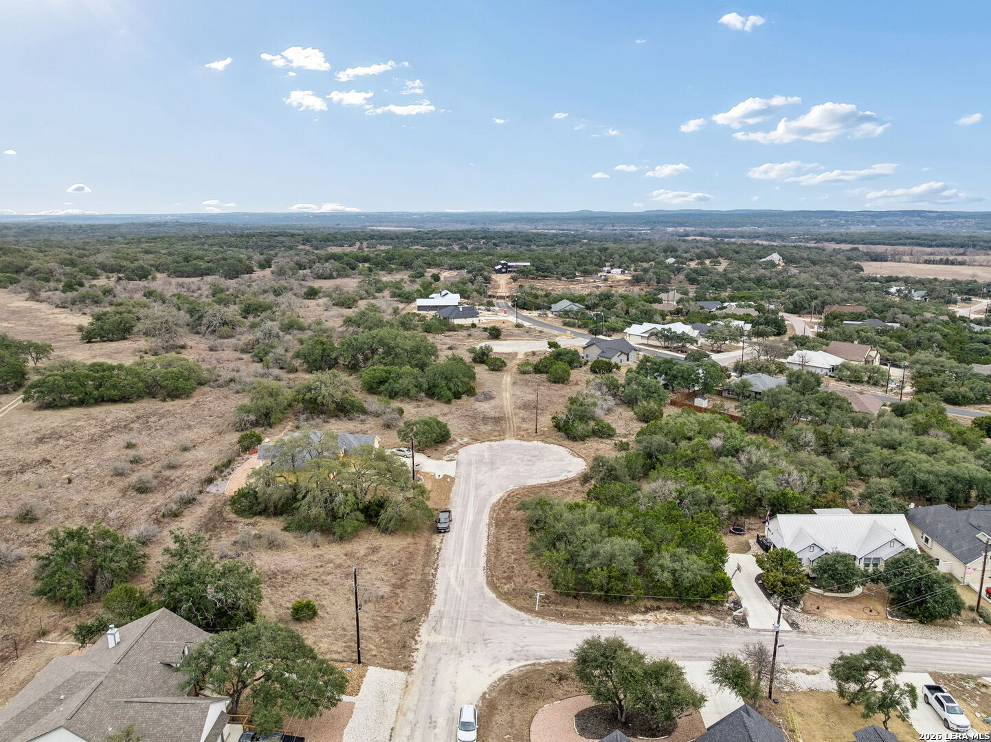 Aerial neighborhood view with dry brush-covered lots, scattered homes, and a curving road.