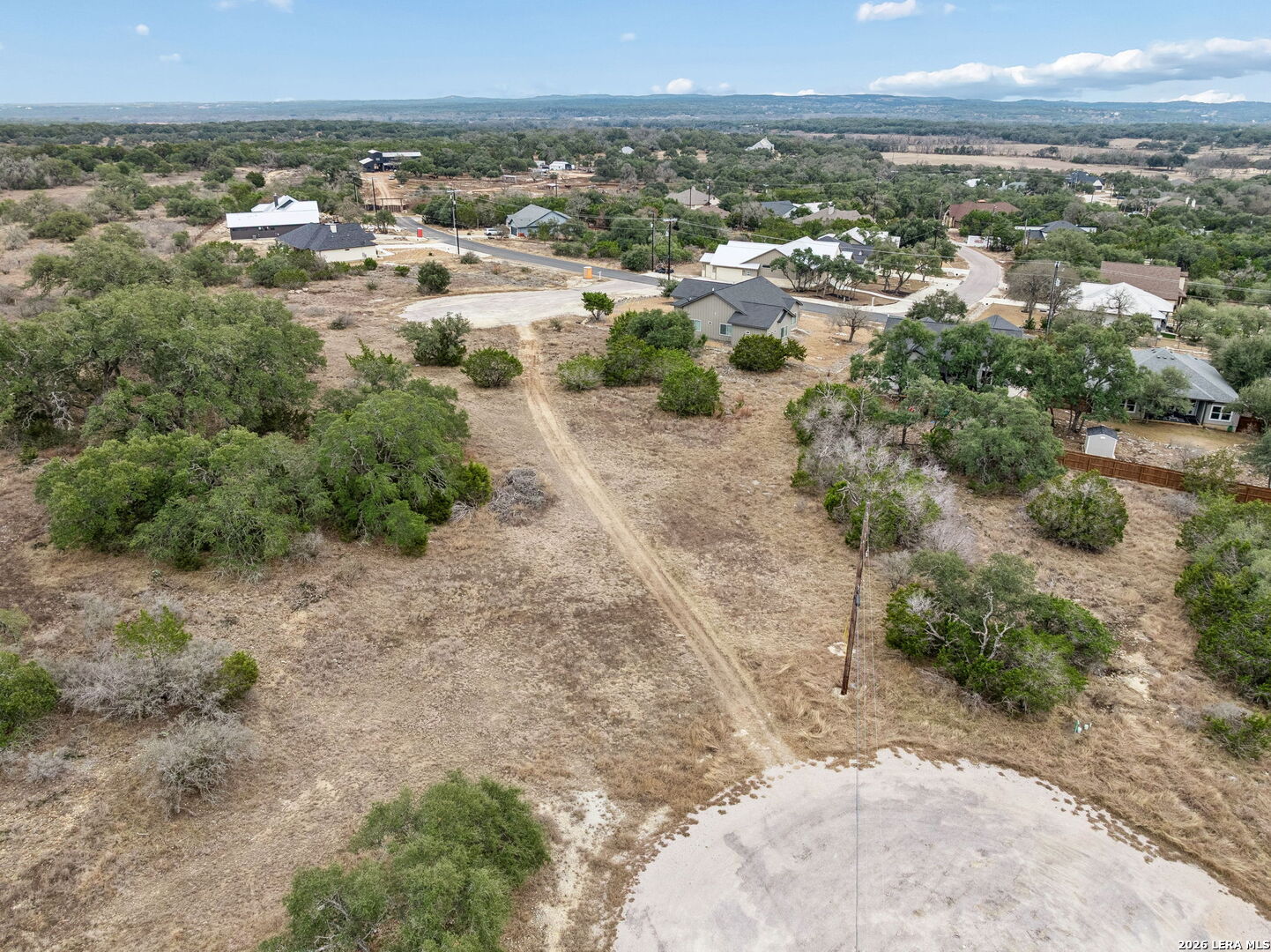 Aerial view of a dry vacant lot with scattered trees, dirt track, and surrounding homes.