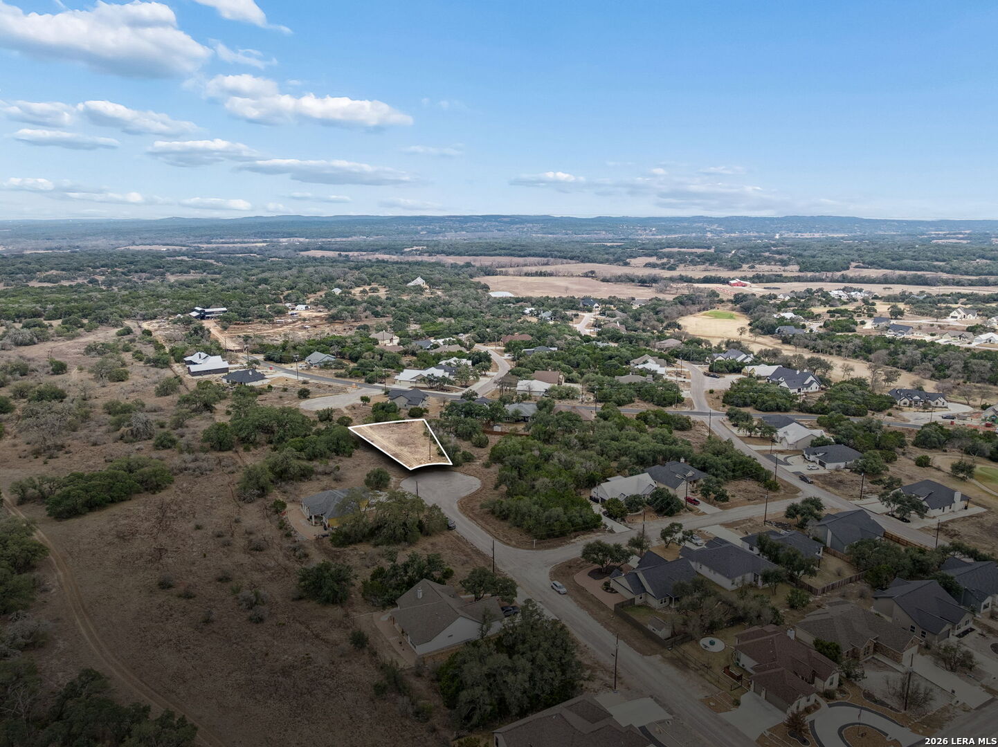 Aerial view of a white-outlined vacant lot in a neighborhood, with rolling wooded hills in the distance.