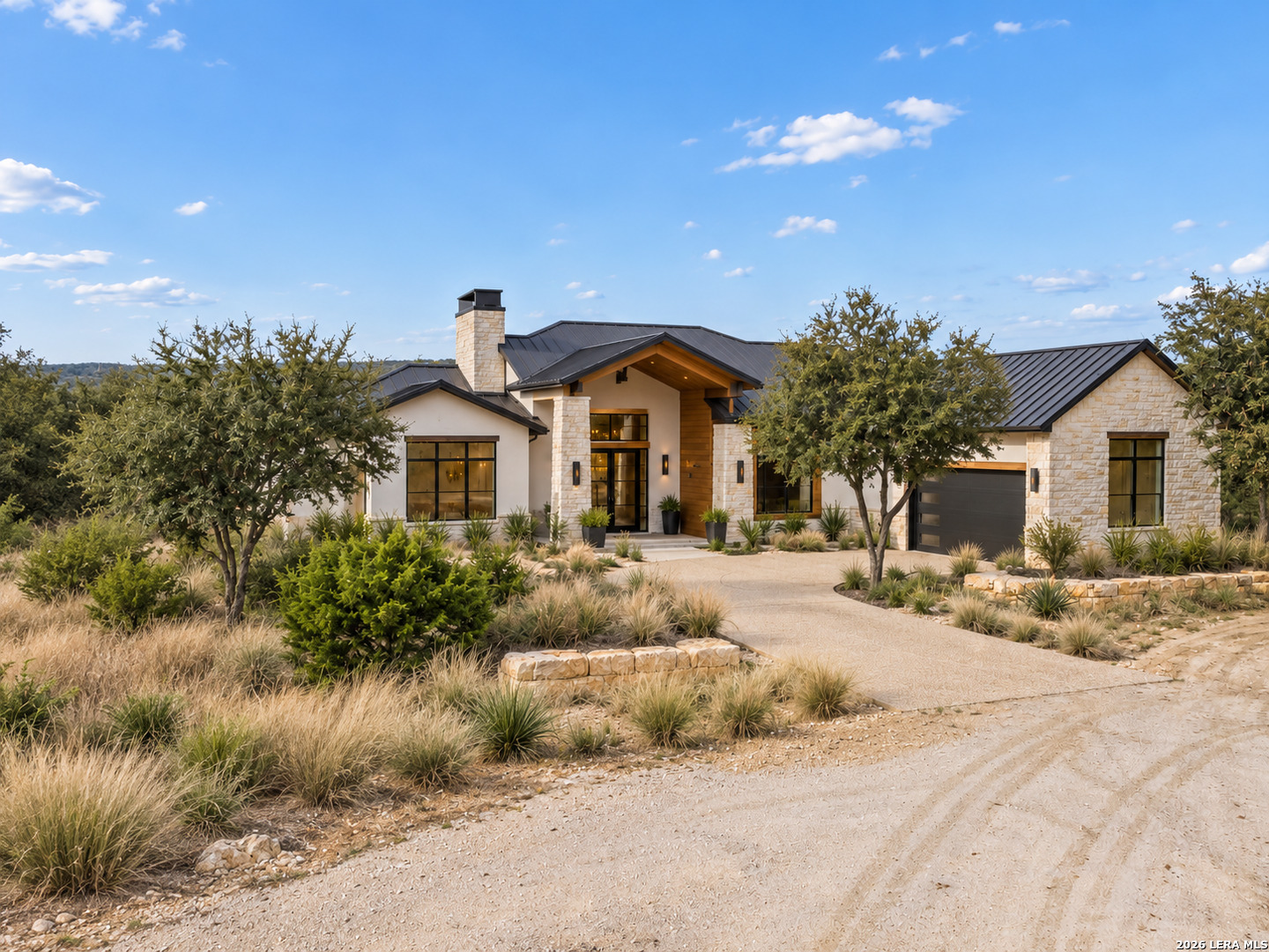 Modern stone-and-stucco home with dark metal roofs, a gravel driveway, and native landscaping.
