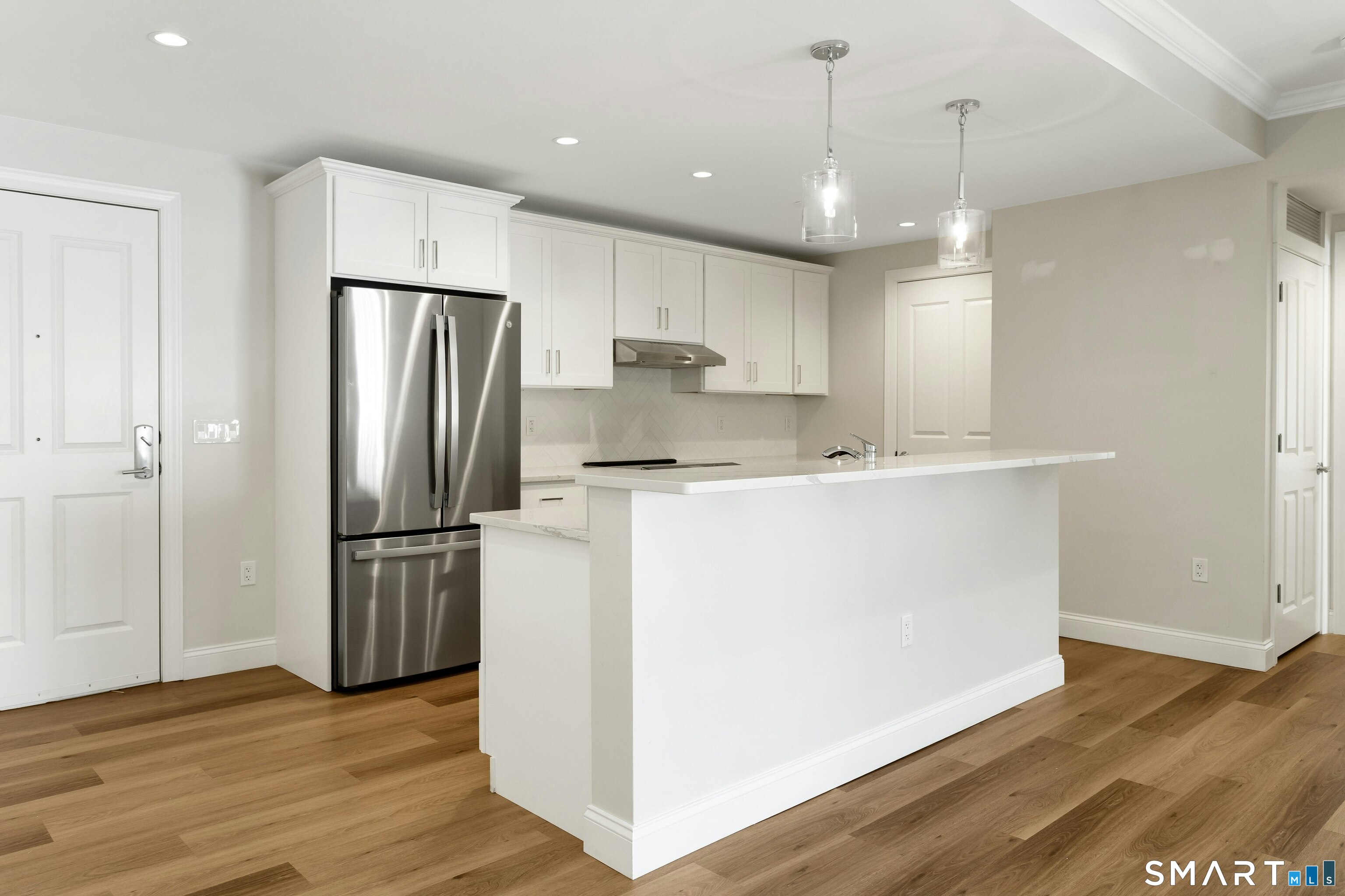 Vacant modern kitchen with a white island, stainless steel refrigerator, and warm wood flooring.