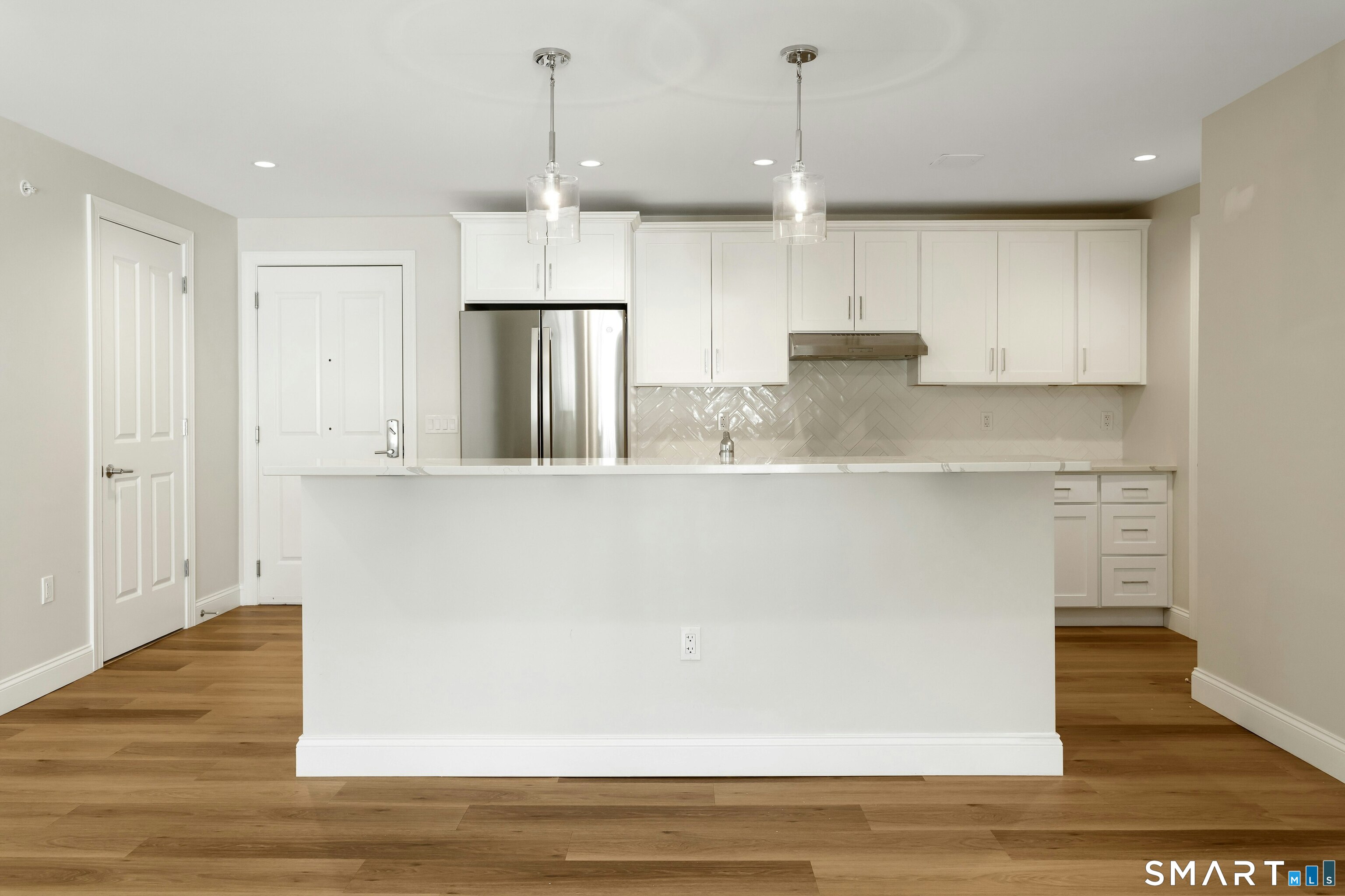 Vacant kitchen with a large white island, stainless steel refrigerator, and glossy herringbone tile backsplash.
