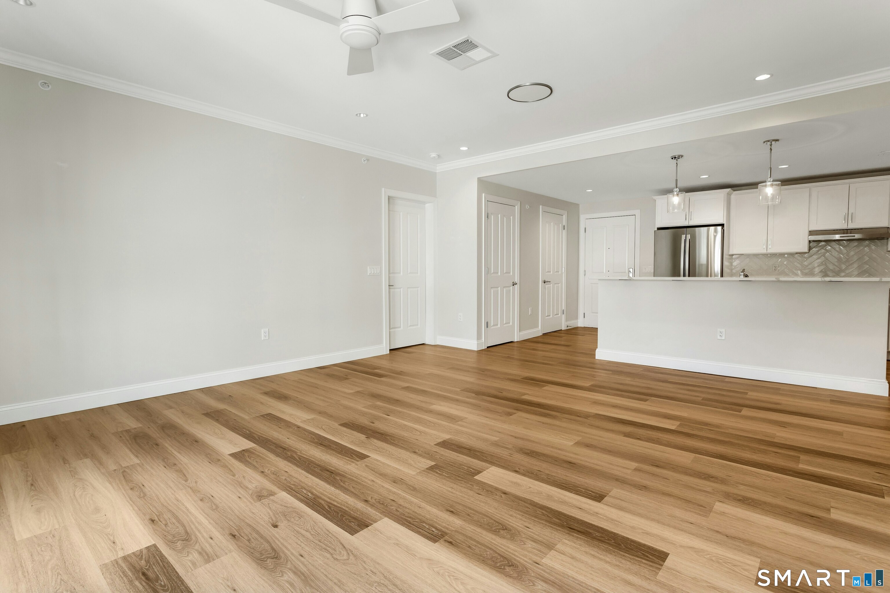 Empty living area opening to a white kitchen, with wide-plank wood flooring and a large peninsula.