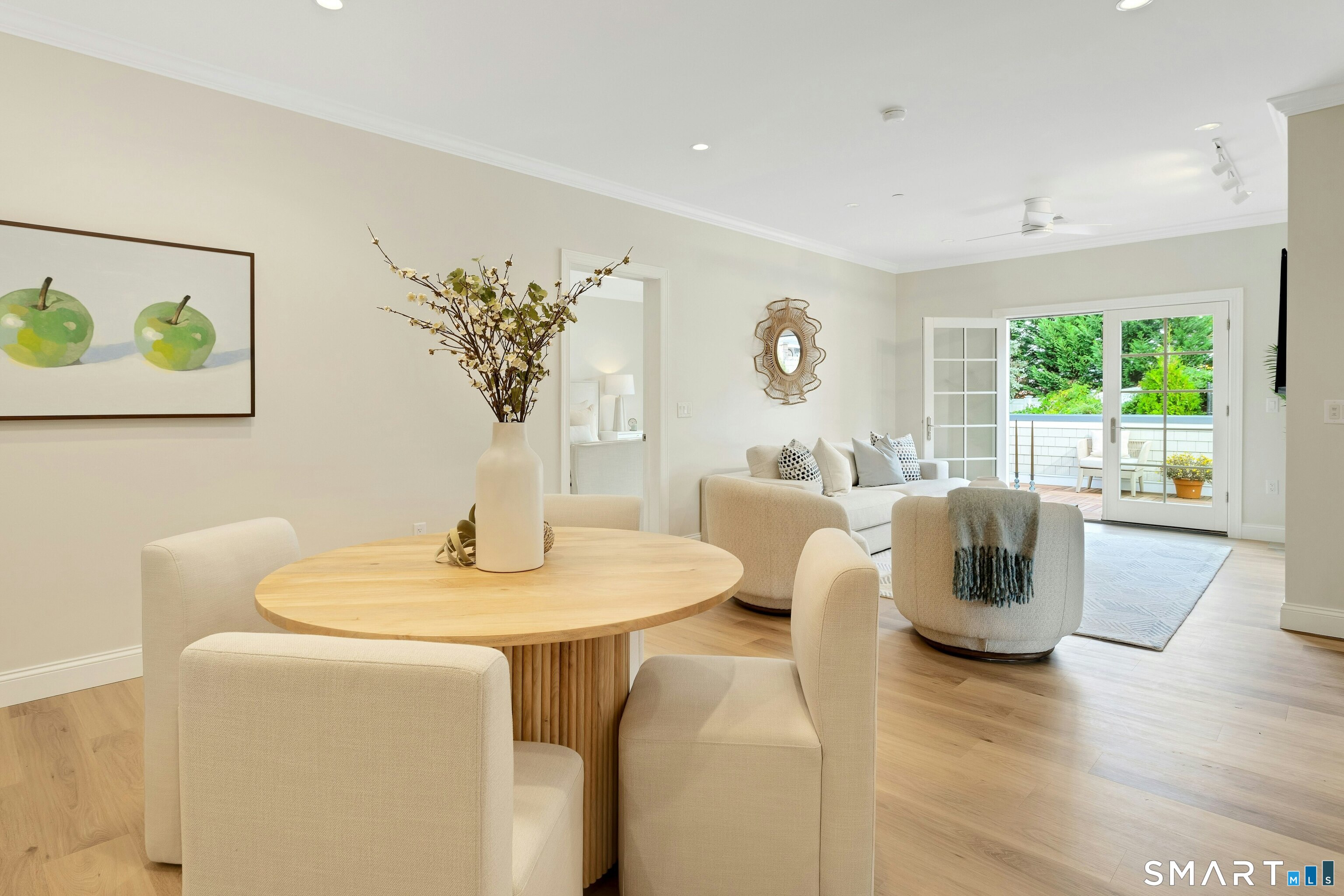 Open-plan living and dining room with a round wood table and glass doors to a leafy balcony.