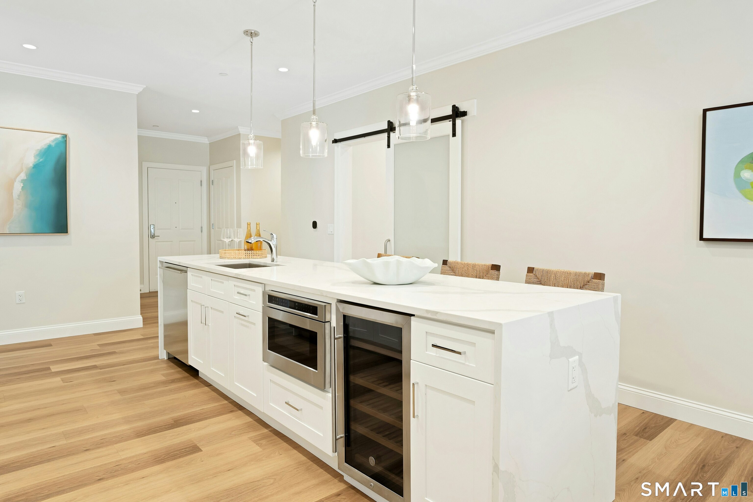 Modern kitchen with a long white island, stainless appliances, wine refrigerator, and pendant lights.