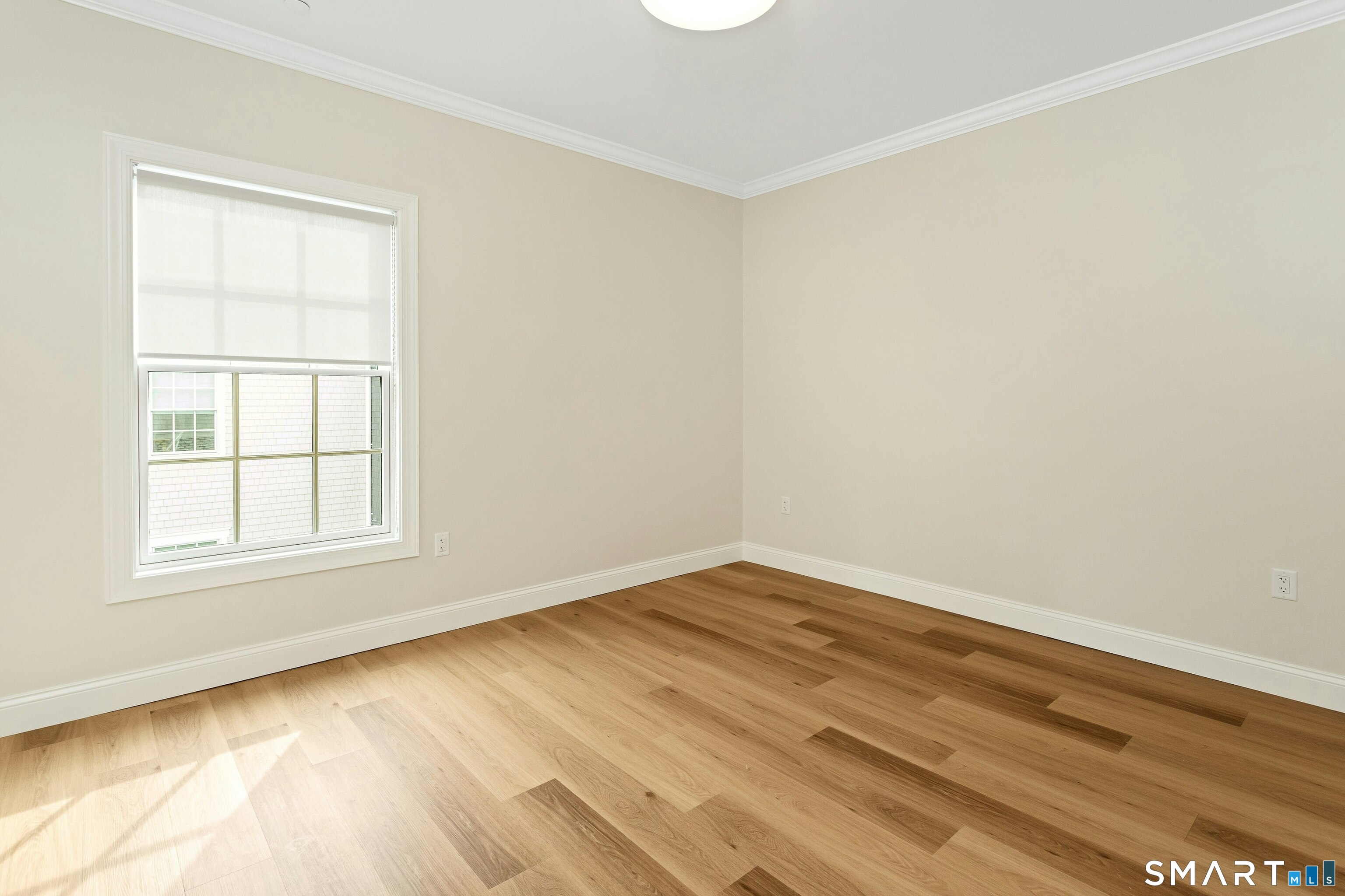 Vacant room with light wood floors and a large shaded window.