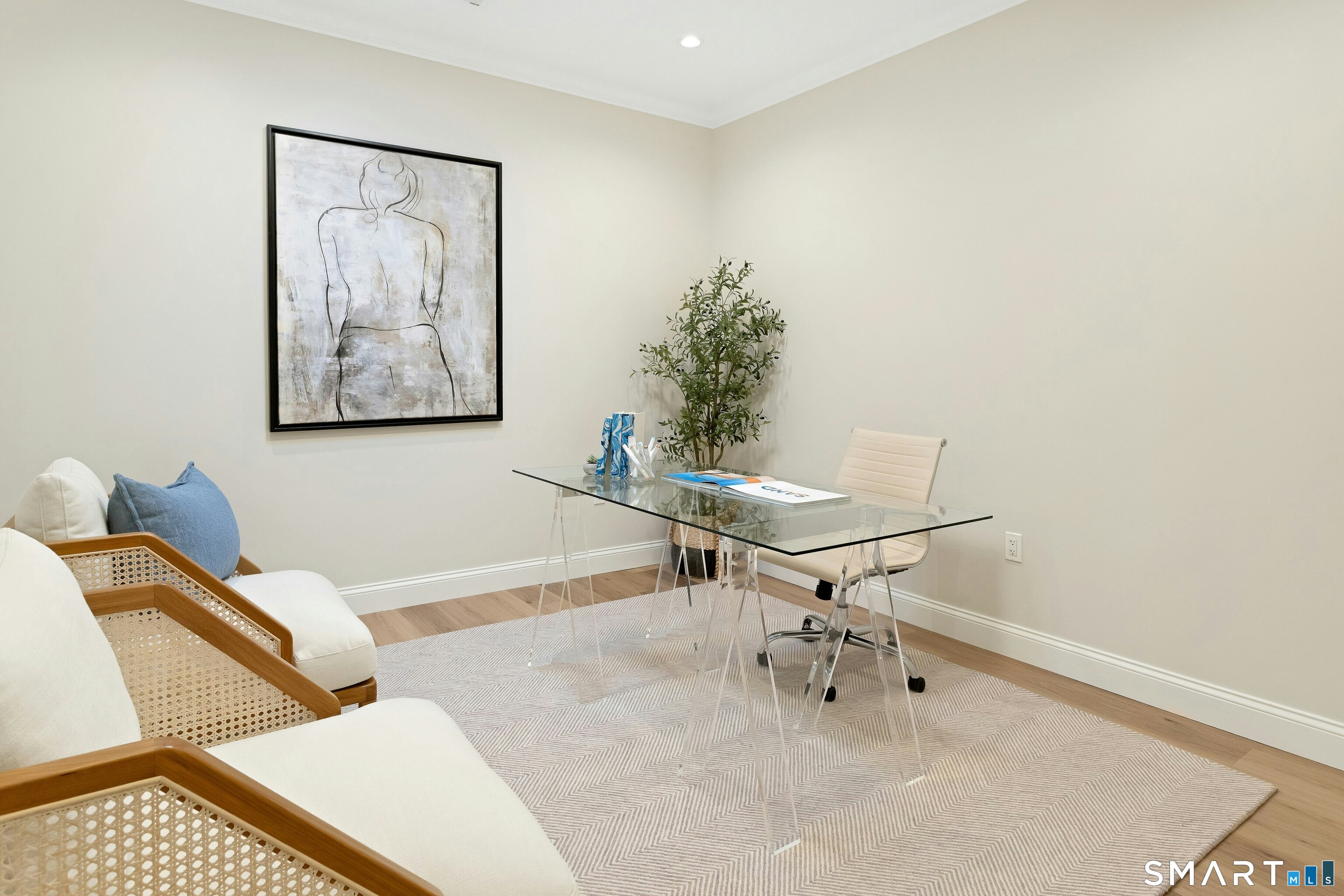 Furnished home office with a glass desk, cane lounge chairs, framed artwork, and a potted tree.