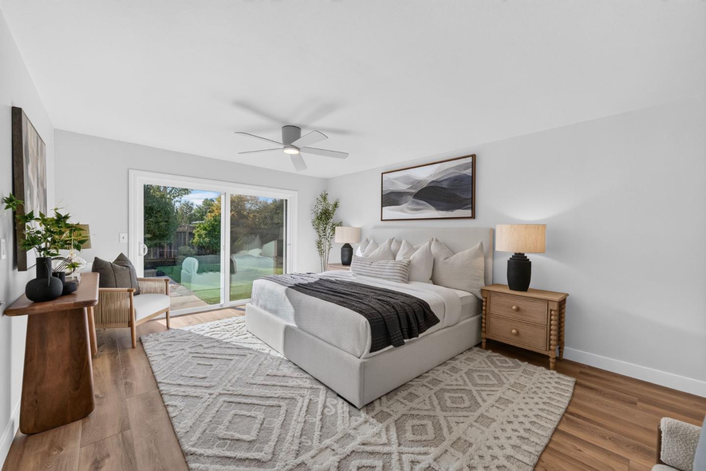 Staged bedroom with a white upholstered bed, patterned rug, and sliding glass doors overlooking a garden.