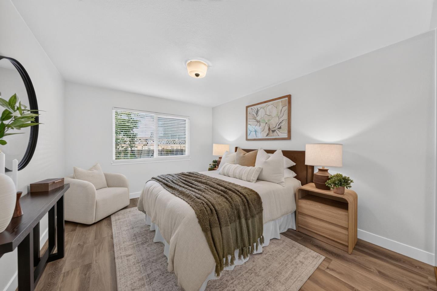 Staged bedroom with a white bed, dark textured throw, wooden nightstand, and window with blinds.