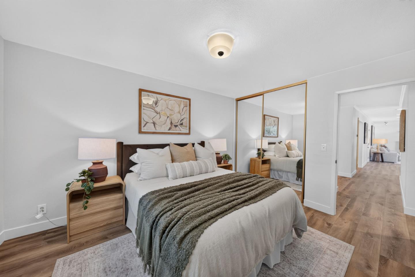 Staged bedroom with a neutral bed, mirrored closet doors, and warm wood flooring.