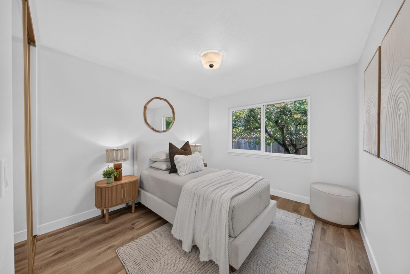 Staged bedroom with a white upholstered bed and large window overlooking leafy trees.