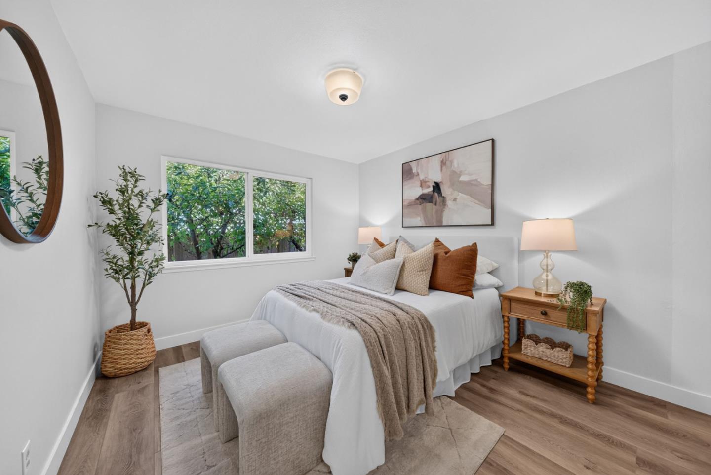 Staged bedroom with a white bed, rust-colored pillows, upholstered benches, and a window overlooking leafy trees.