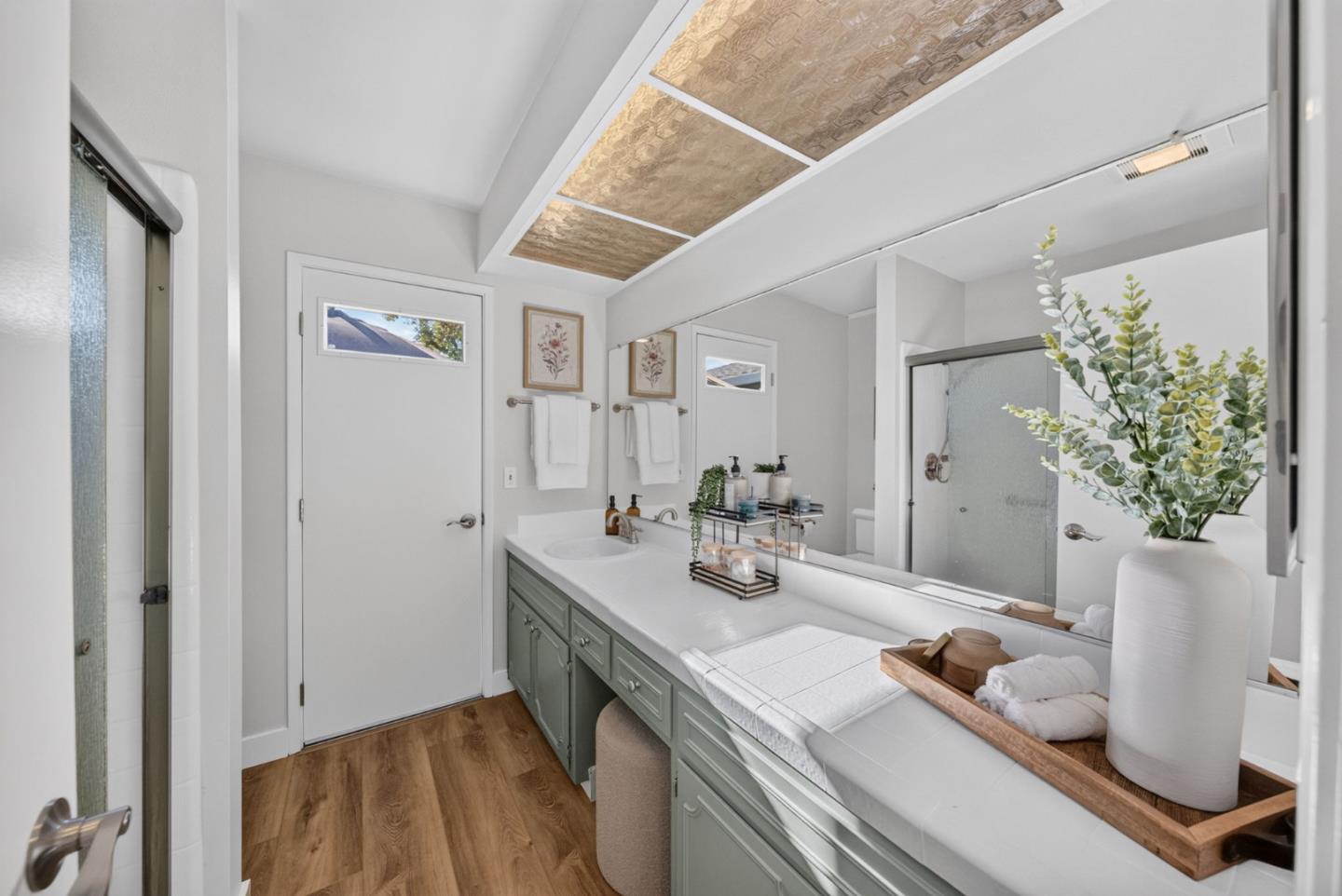 Bathroom with a sage-green vanity, long mirror, glass shower, and wood flooring.