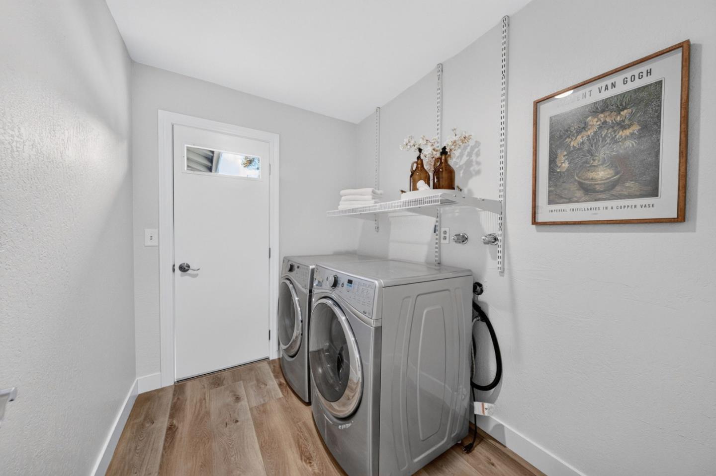 Laundry room with front-loading washer and dryer, wire shelving, and framed floral artwork.