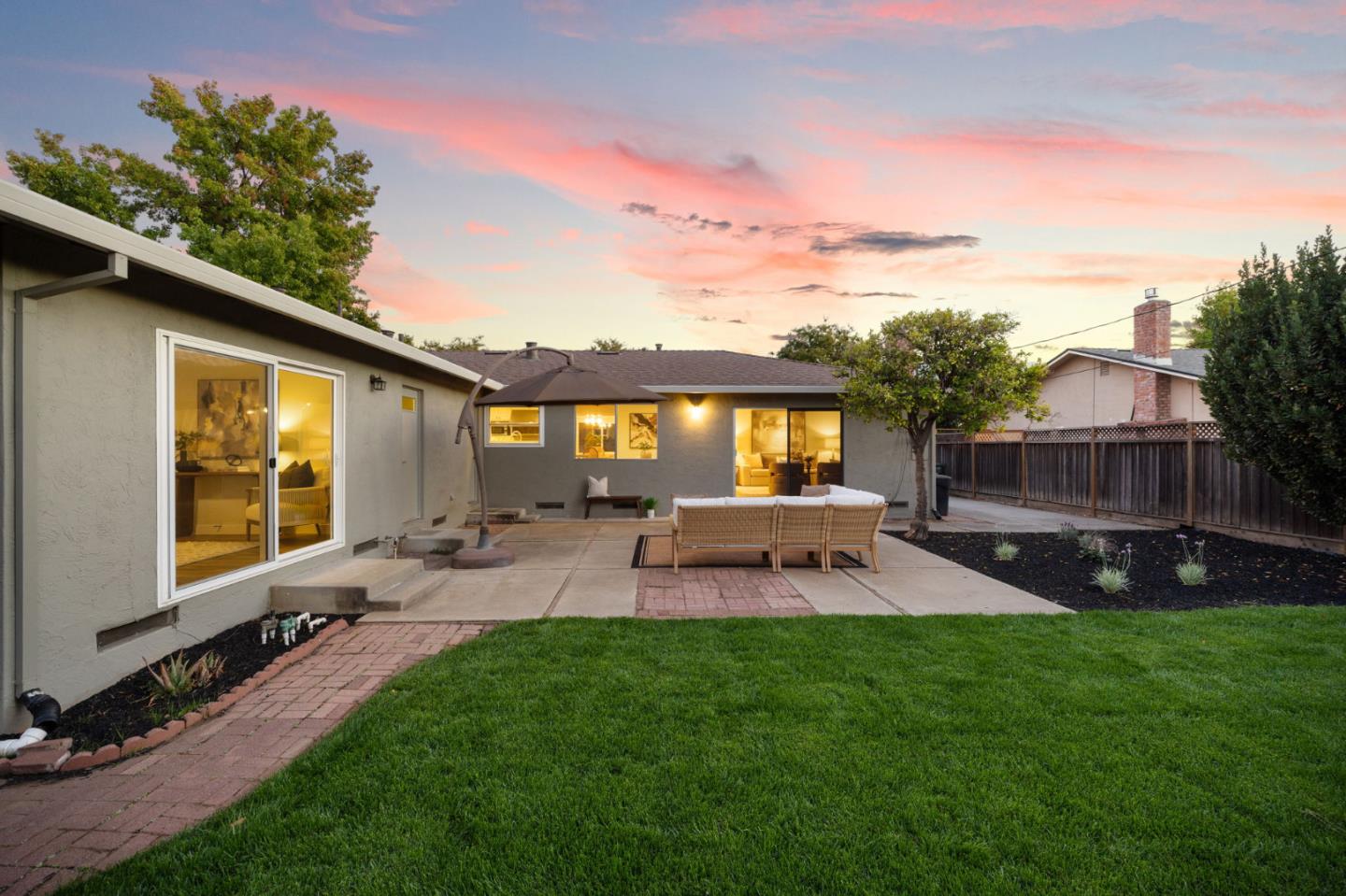 Home’s rear patio and lawn at dusk, with outdoor dining furniture and warm light glowing through the windows.