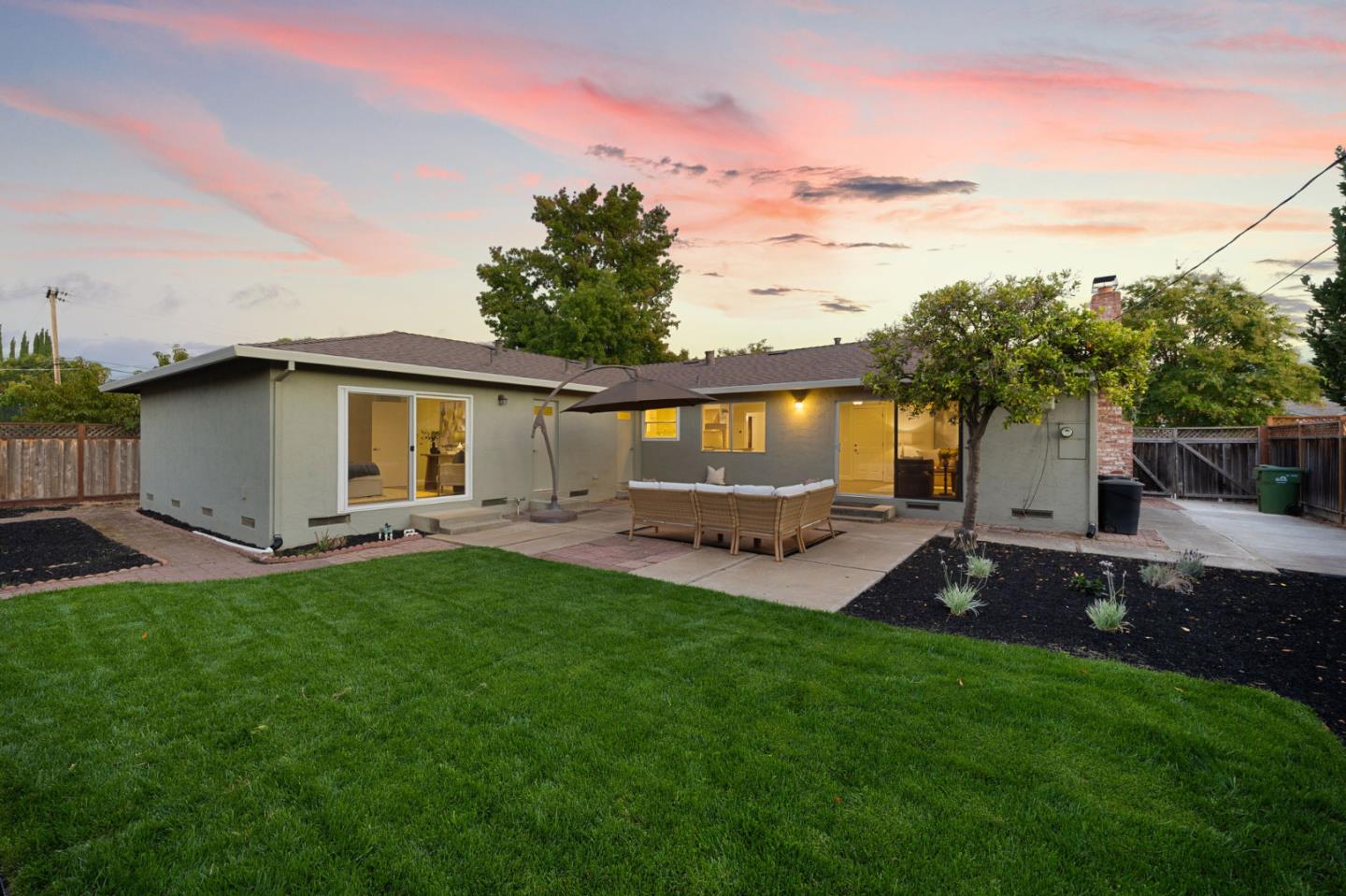 Single-story home and backyard patio with outdoor seating beneath a pink dusk sky.