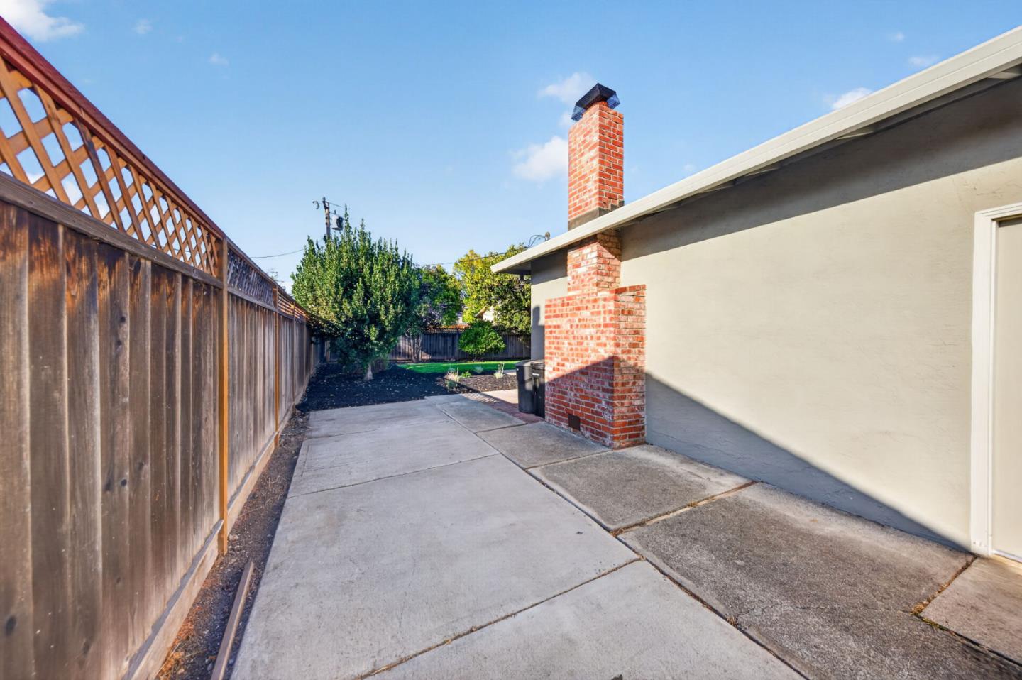 Concrete side yard beside a house, bordered by a tall wooden fence and featuring a brick chimney.
