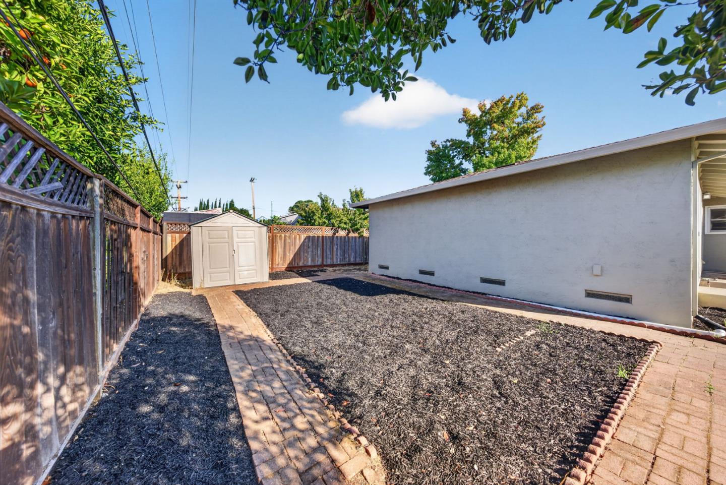 Fenced backyard with mulched garden beds, brick paths, a small shed, and the house wall.