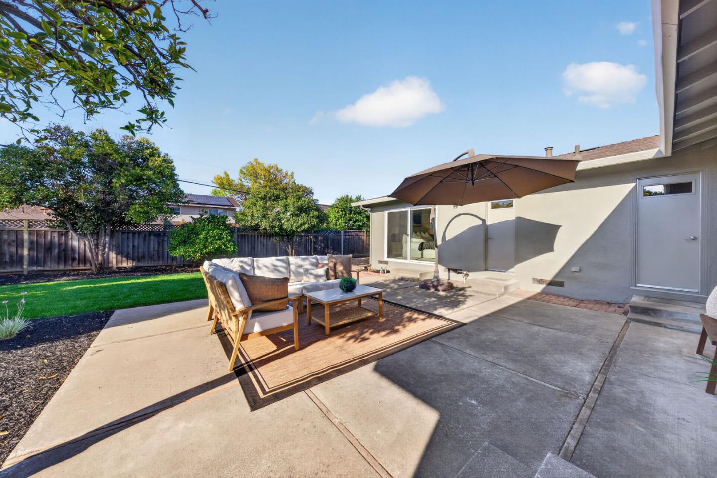 Backyard patio with cushioned seating, a large umbrella, and a lawn bordered by trees and fencing.