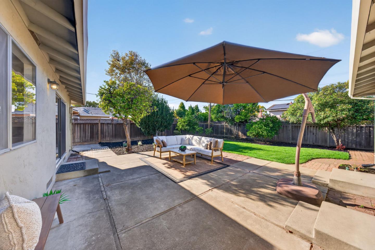 Backyard patio with a large shade umbrella, white outdoor sofa, paved seating area, and fenced lawn.