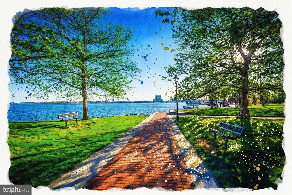 Waterfront park with a brick path, leafy trees, benches, and boats in the distance.