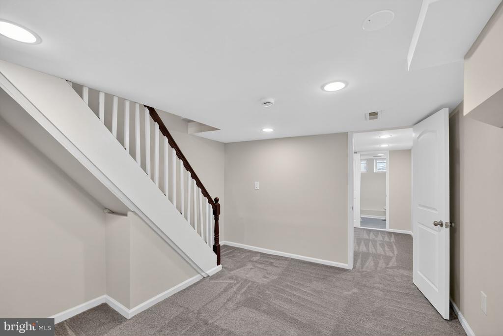 Empty carpeted basement room with a white staircase and an open doorway to another finished area.