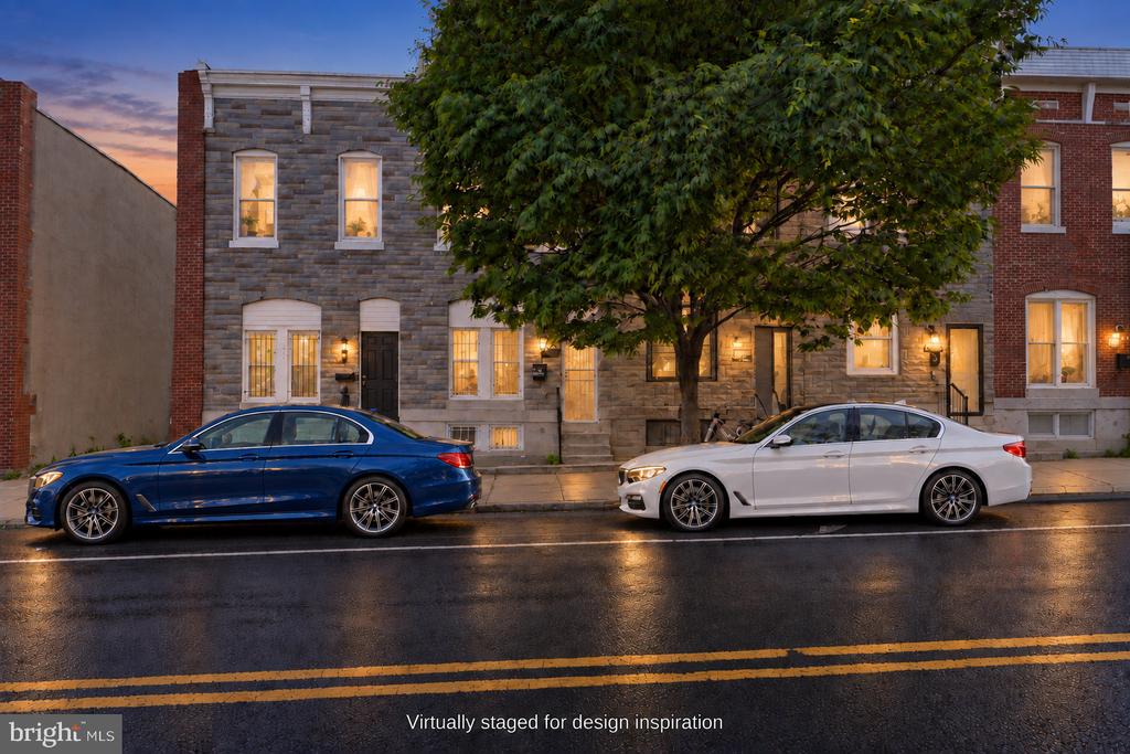 Stone-front Baltimore rowhouse at dusk, partly shaded by a large tree, with cars parked on the wet street.