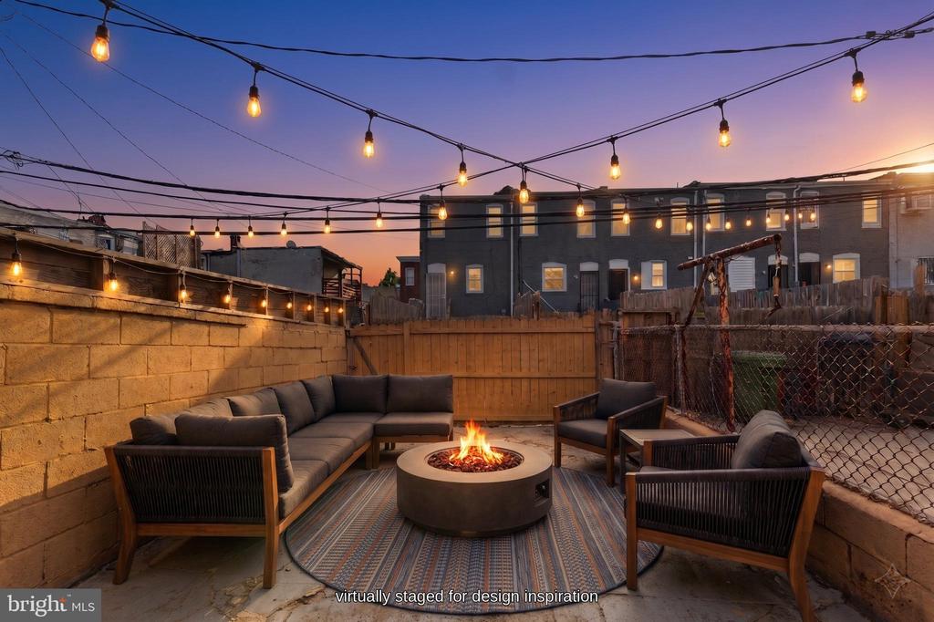 Backyard patio with lounge seating around a fire pit beneath glowing string lights at dusk.