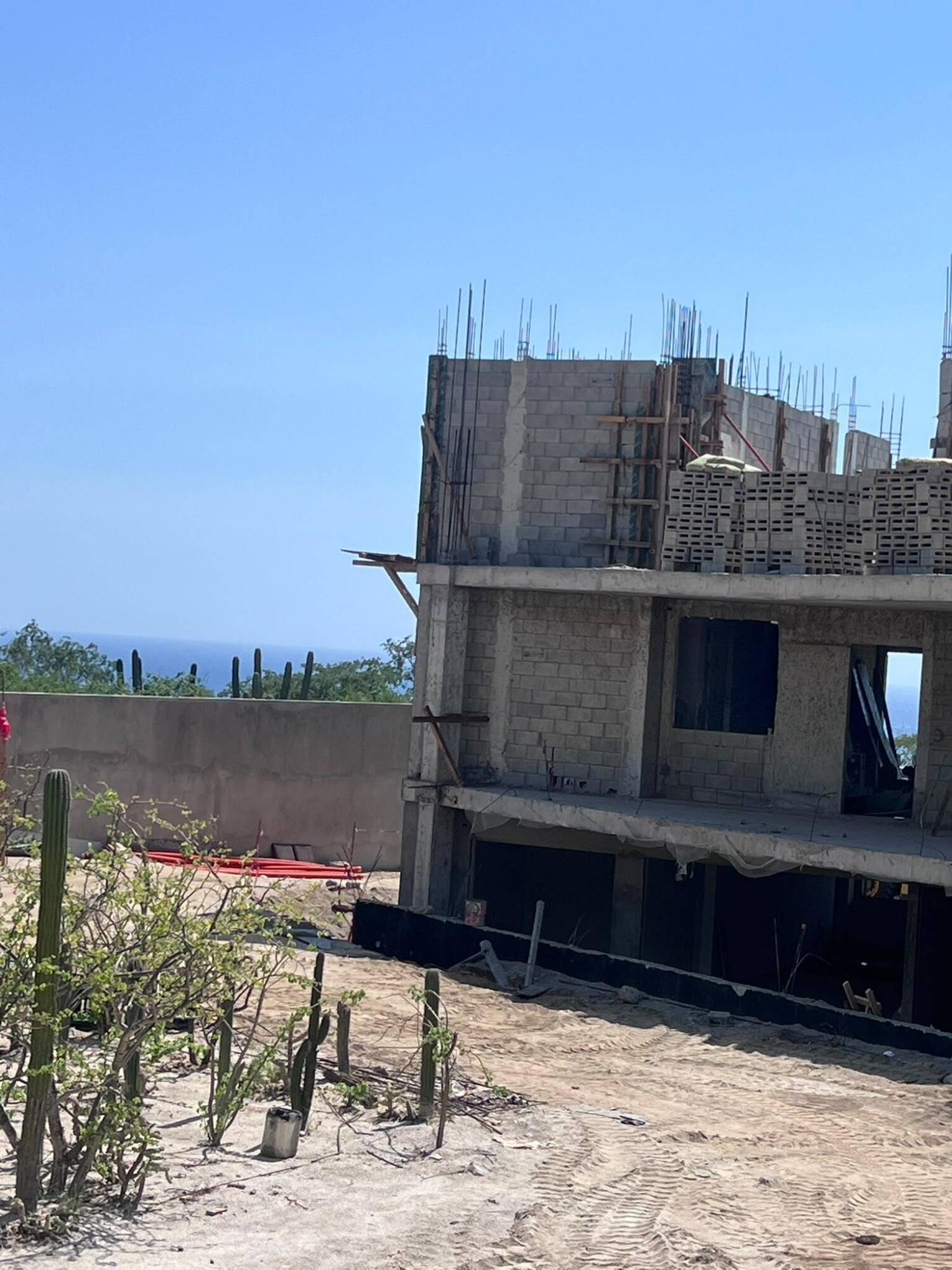Unfinished multi-story building with exposed rebar, cacti in the sandy foreground, and ocean beyond.
