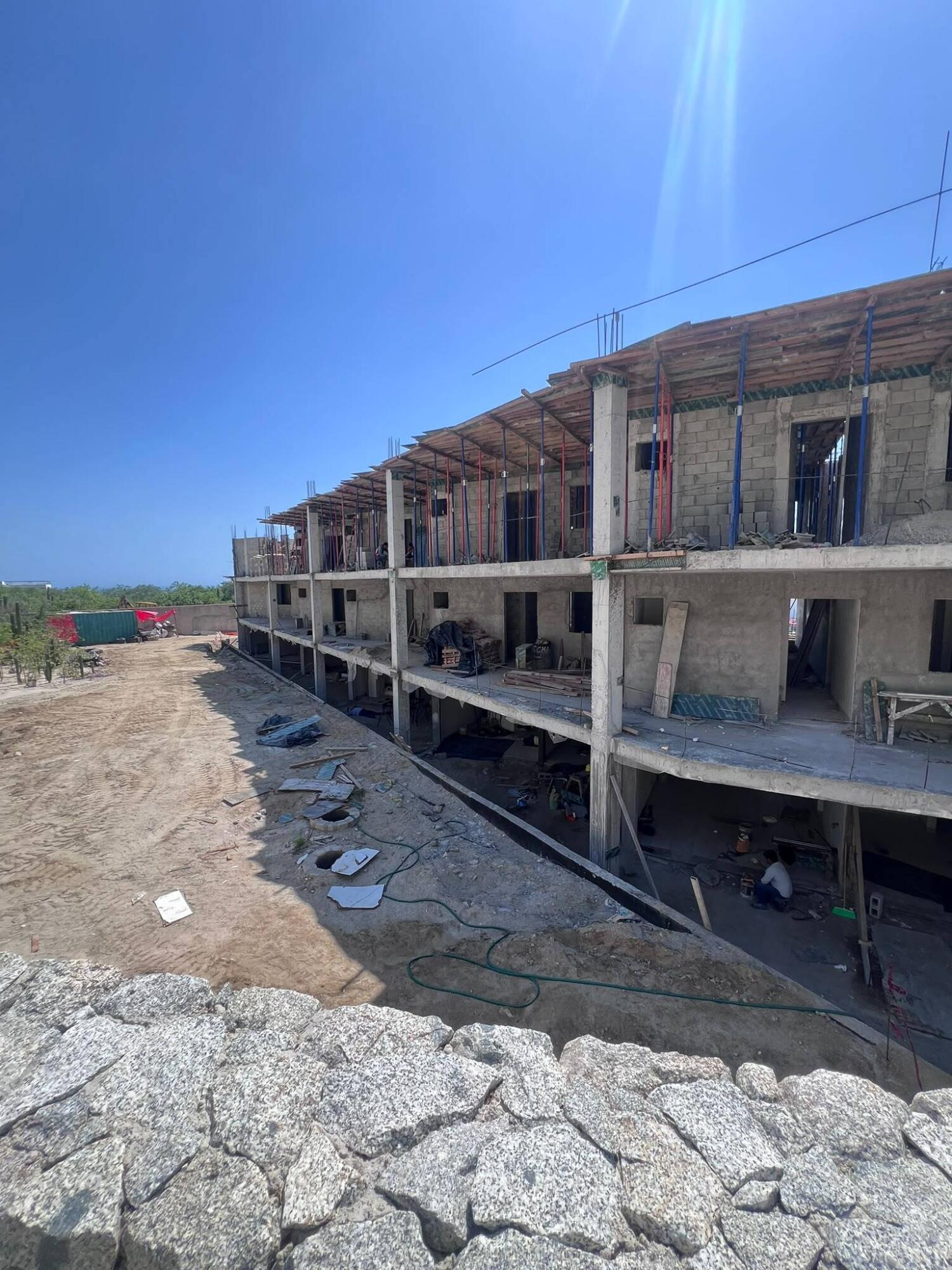 Multi-story building under construction, with exposed concrete, construction supports, and a stone retaining wall.