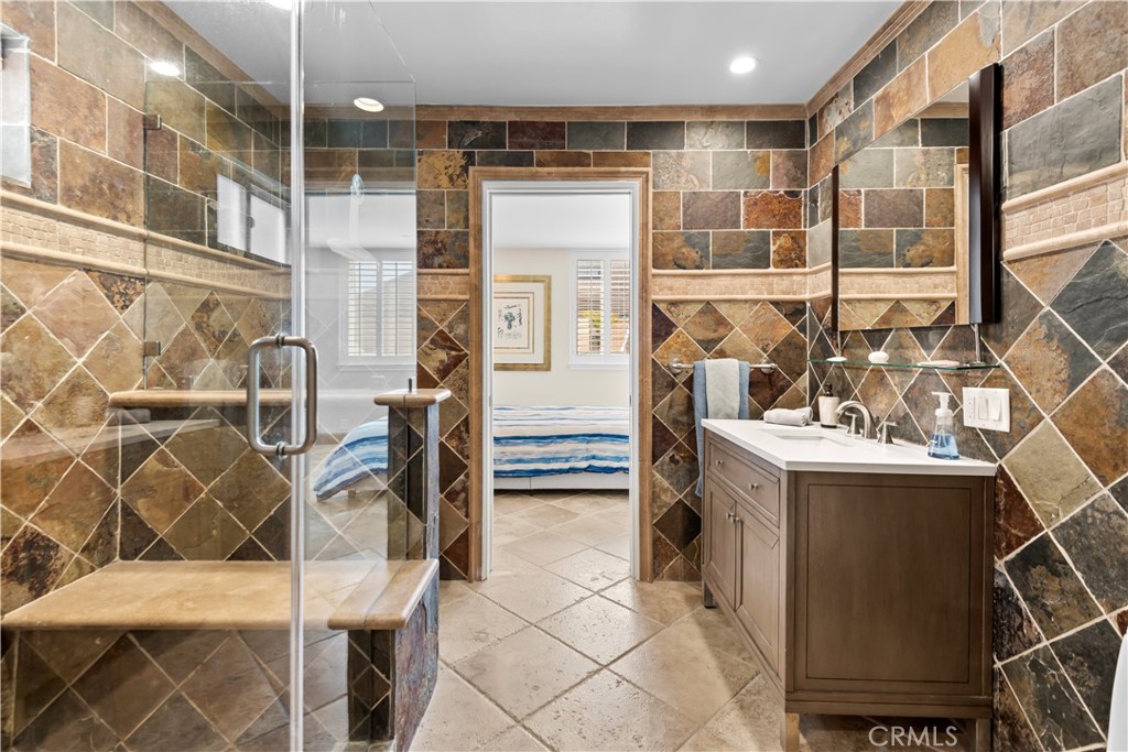 Bathroom with a glass shower, multicolored stone-pattern tile, and a wood vanity beside a bedroom doorway.
