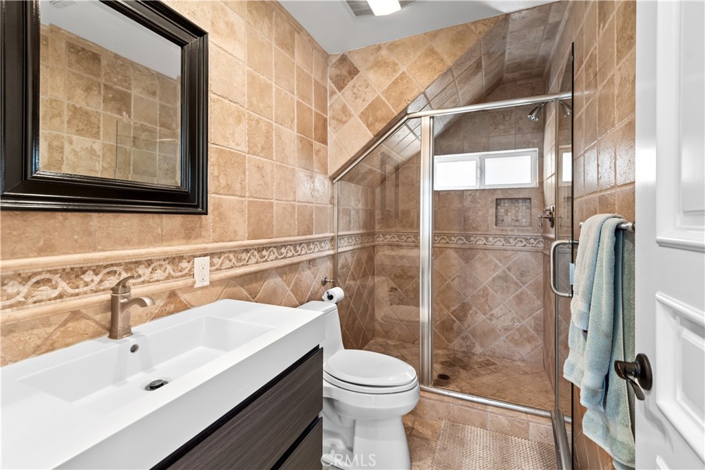 Tan-tiled bathroom with a glass shower, white vanity sink, and blue towels.