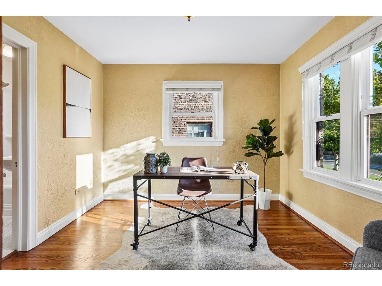 Staged home office with a rolling desk, gray rug, and large sunlit windows.