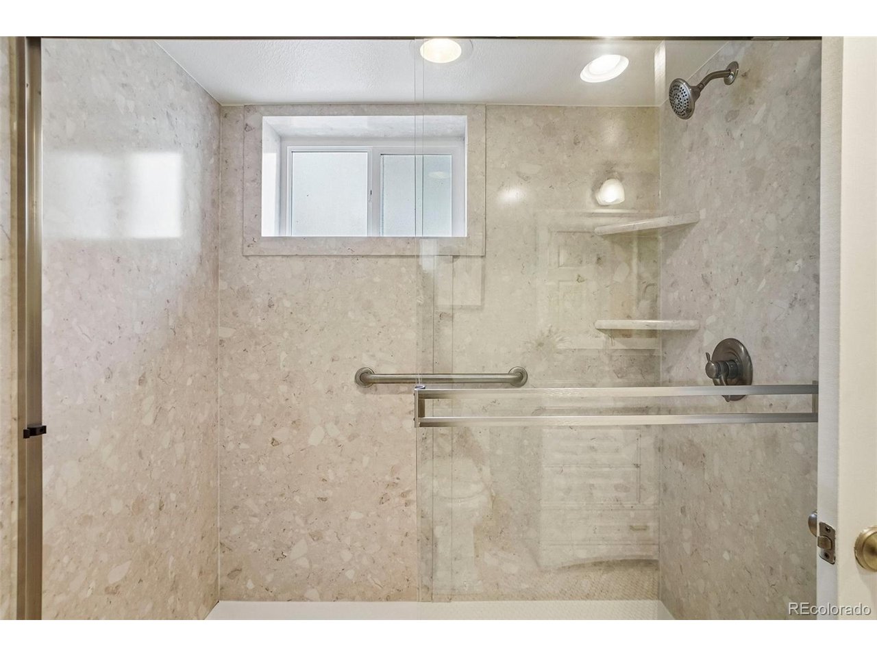 Walk-in shower with beige stone tile, glass enclosure, grab bar, and built-in corner shelves.