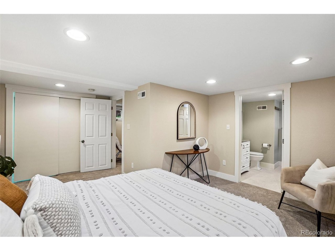 Furnished bedroom with a white patterned bedspread, arched mirror, and adjoining bathroom.