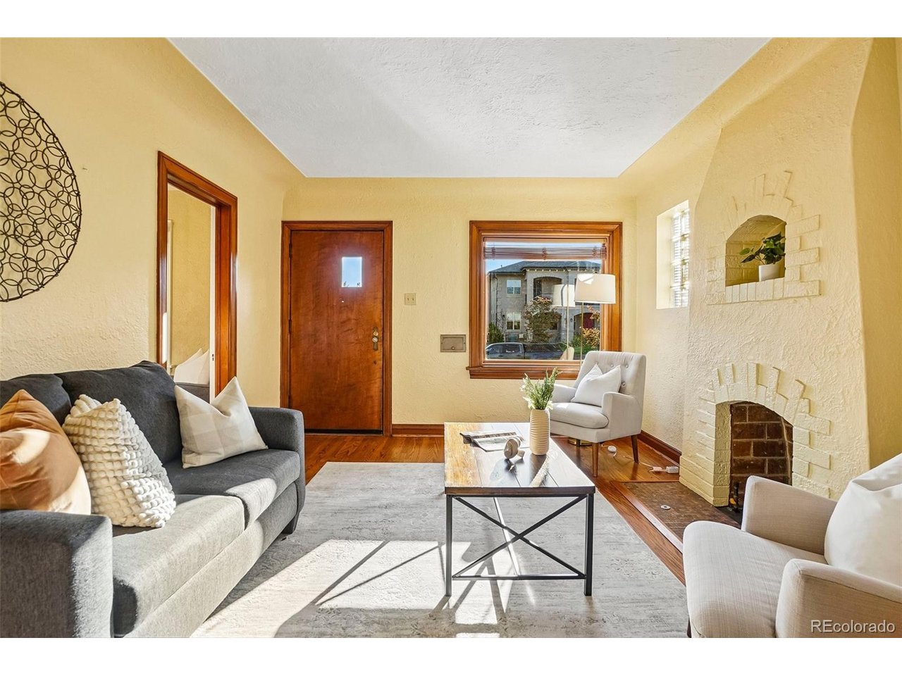 Staged living room with a white brick fireplace, gray sofa, hardwood floors, and a wood-trimmed front door.