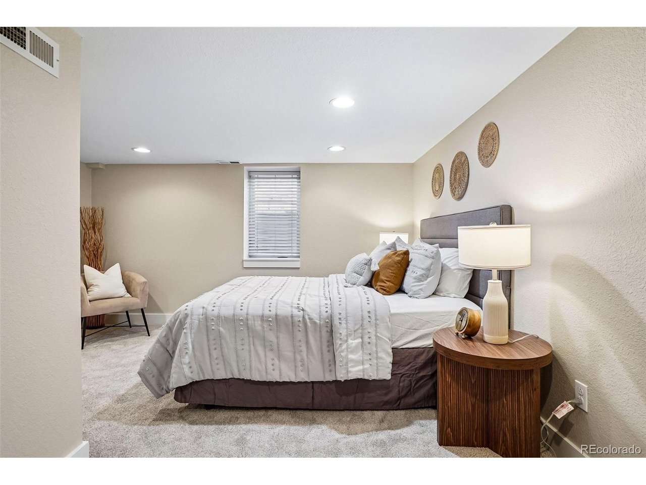 Basement bedroom with a gray upholstered bed, patterned white bedding, and a round wooden nightstand.