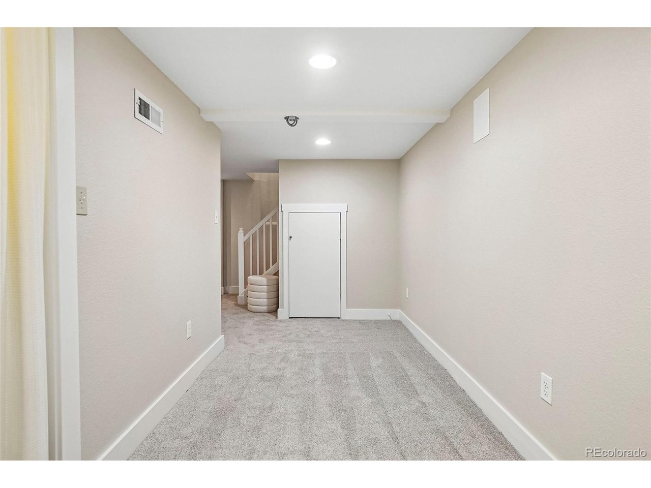 Carpeted basement room with beige walls, a small storage door, and stairs at the far end.