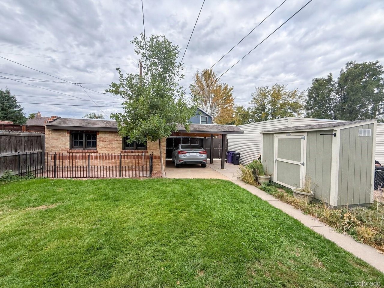 Brick home and covered carport beyond a grassy backyard, with a detached shed and concrete path.