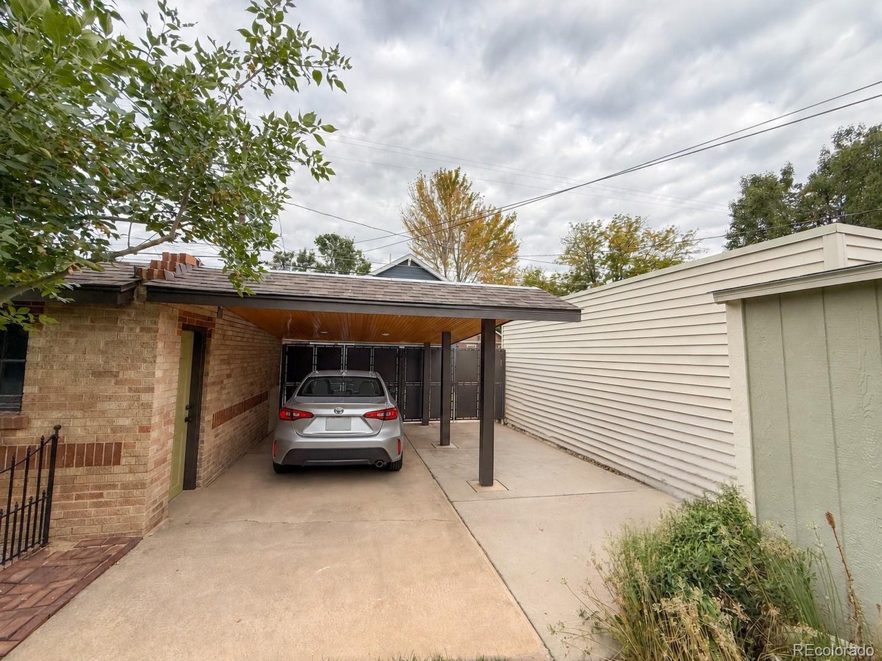 Covered carport beside a brick house, with a silver car and tall privacy fence.
