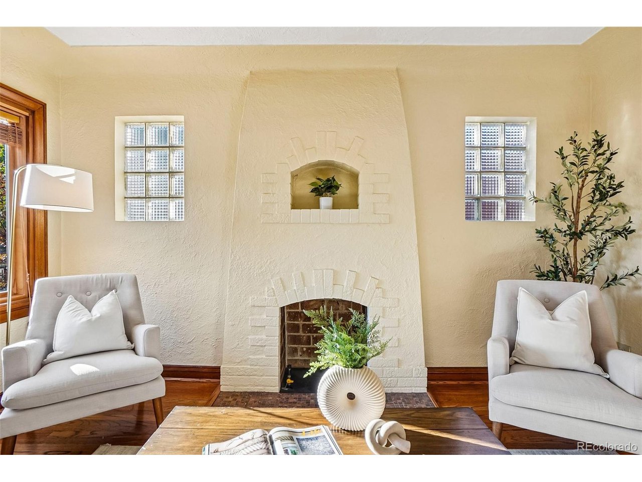 Staged living room with a cream fireplace, glass-block windows, and two light upholstered armchairs.
