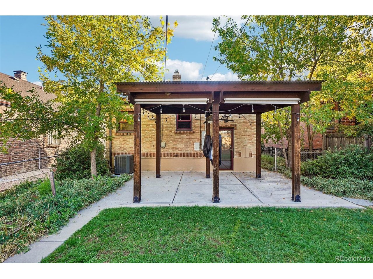 Brick home’s rear patio beneath a wooden pergola with string lights, bordered by lawn and mature trees.