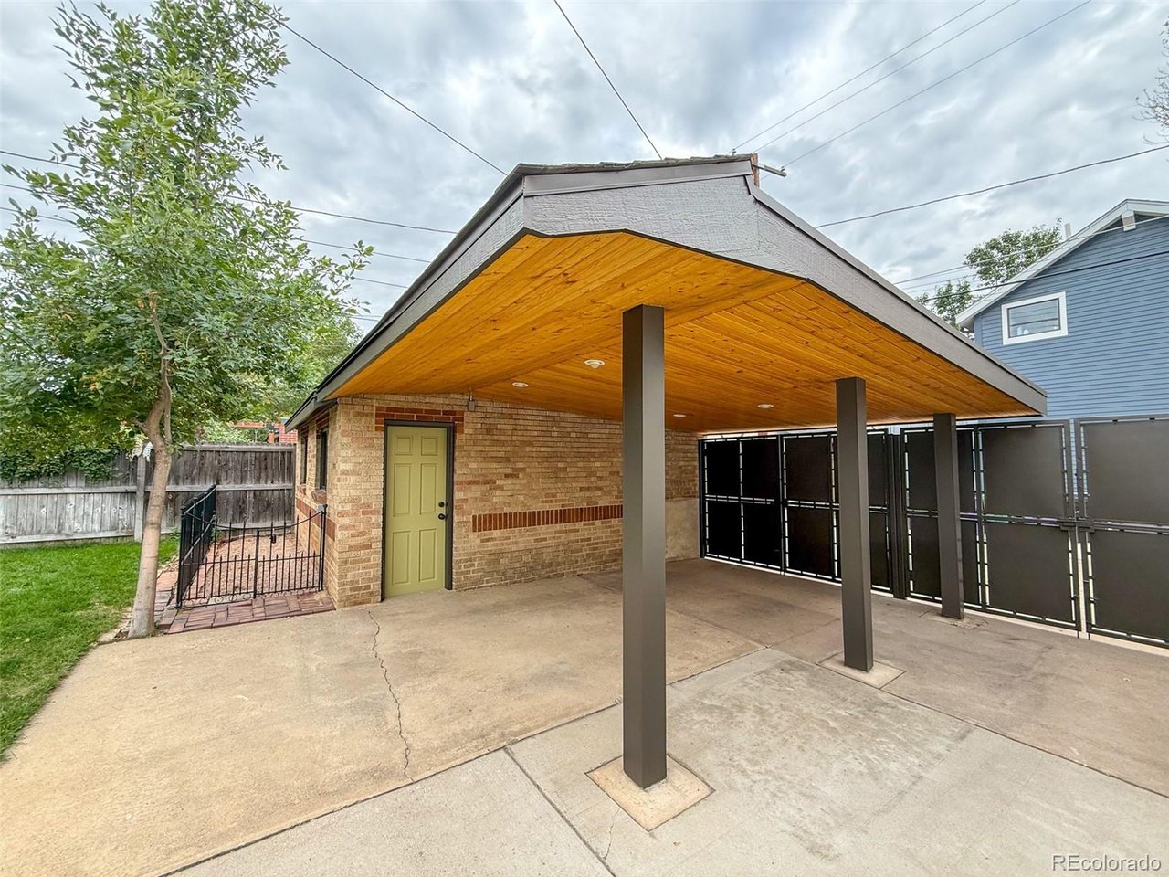 Covered patio with a wood-plank ceiling, dark posts, and gated enclosure beside a brick home.