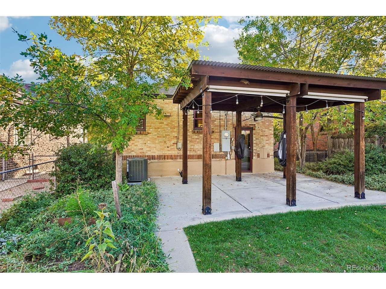 Brick home’s covered patio with timber supports and string lights beside a leafy yard.