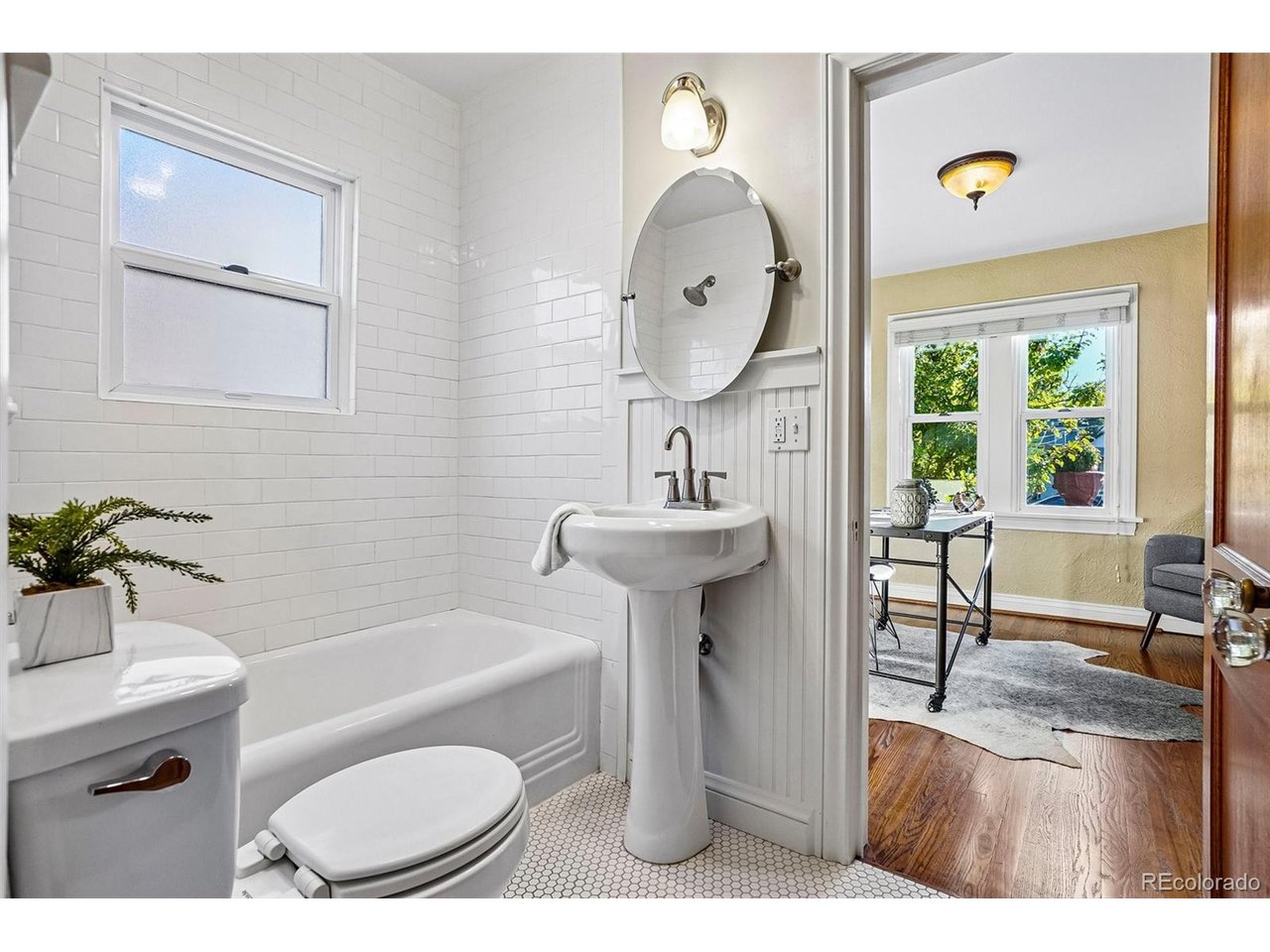 White-tiled bathroom with a bathtub and pedestal sink, opening onto a sunlit room.