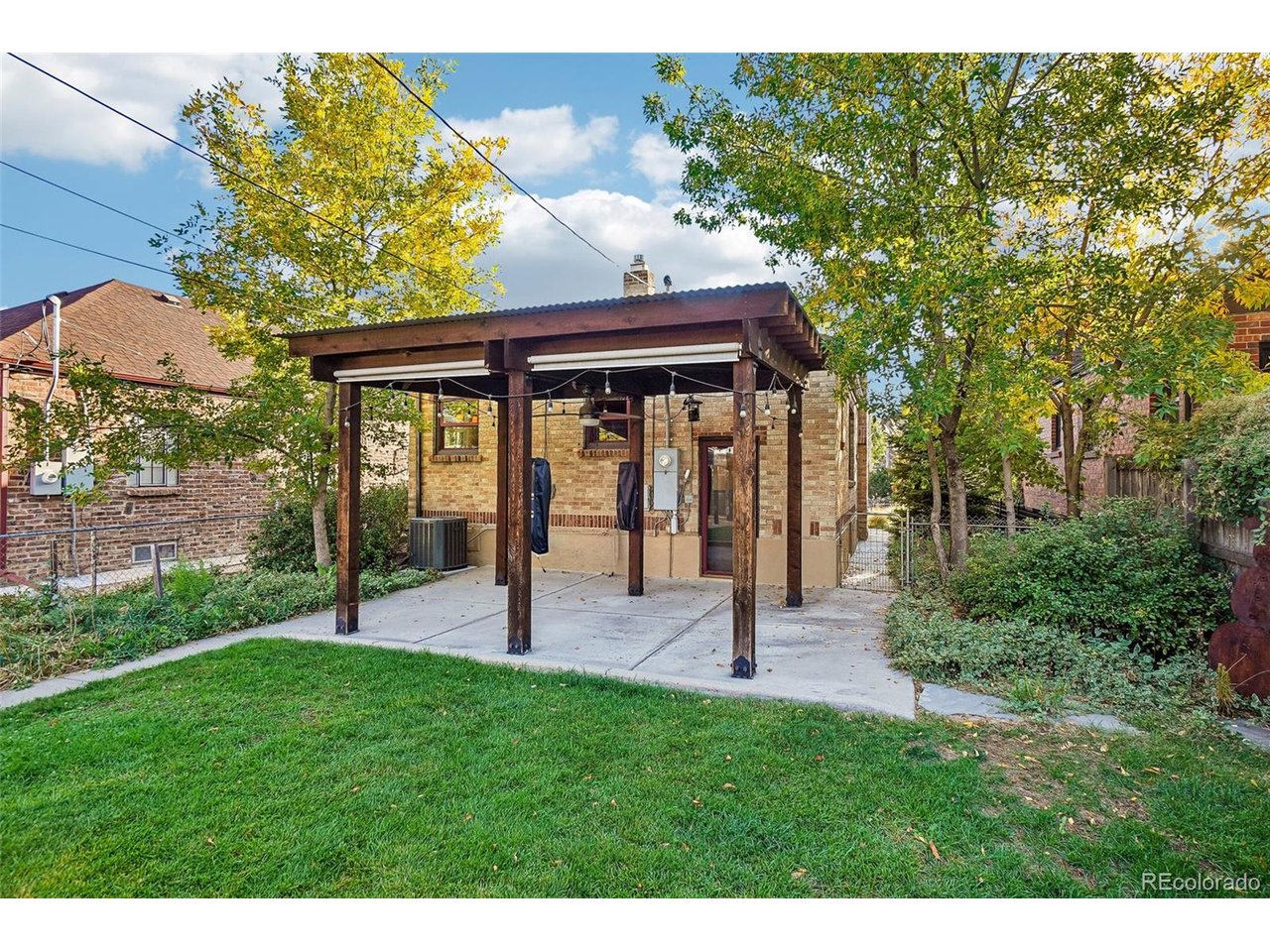 Backyard patio with a dark wood pergola beside a brick house, surrounded by lawn and mature trees.