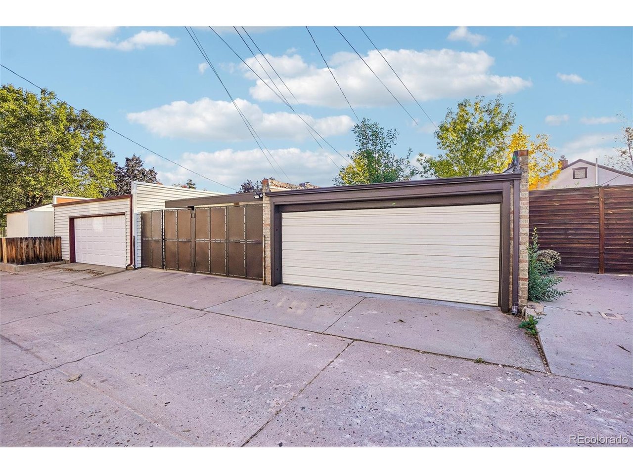 Detached garages with white doors line a paved alley beside wood fencing.