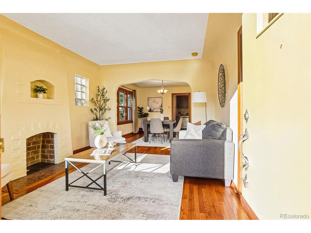 Staged living room with a white brick fireplace, gray sofa, and arched opening to the dining area.