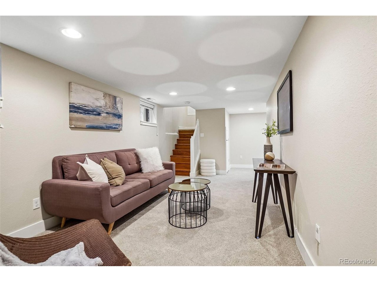 Basement living room with a mauve sofa, round nesting tables, and wooden stairs.