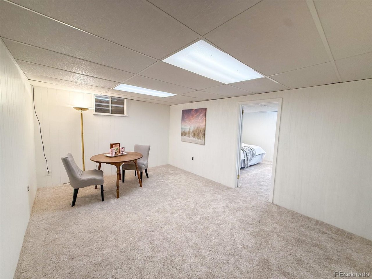 Furnished basement room with beige carpet, drop ceiling, small table, and doorway to a bedroom.